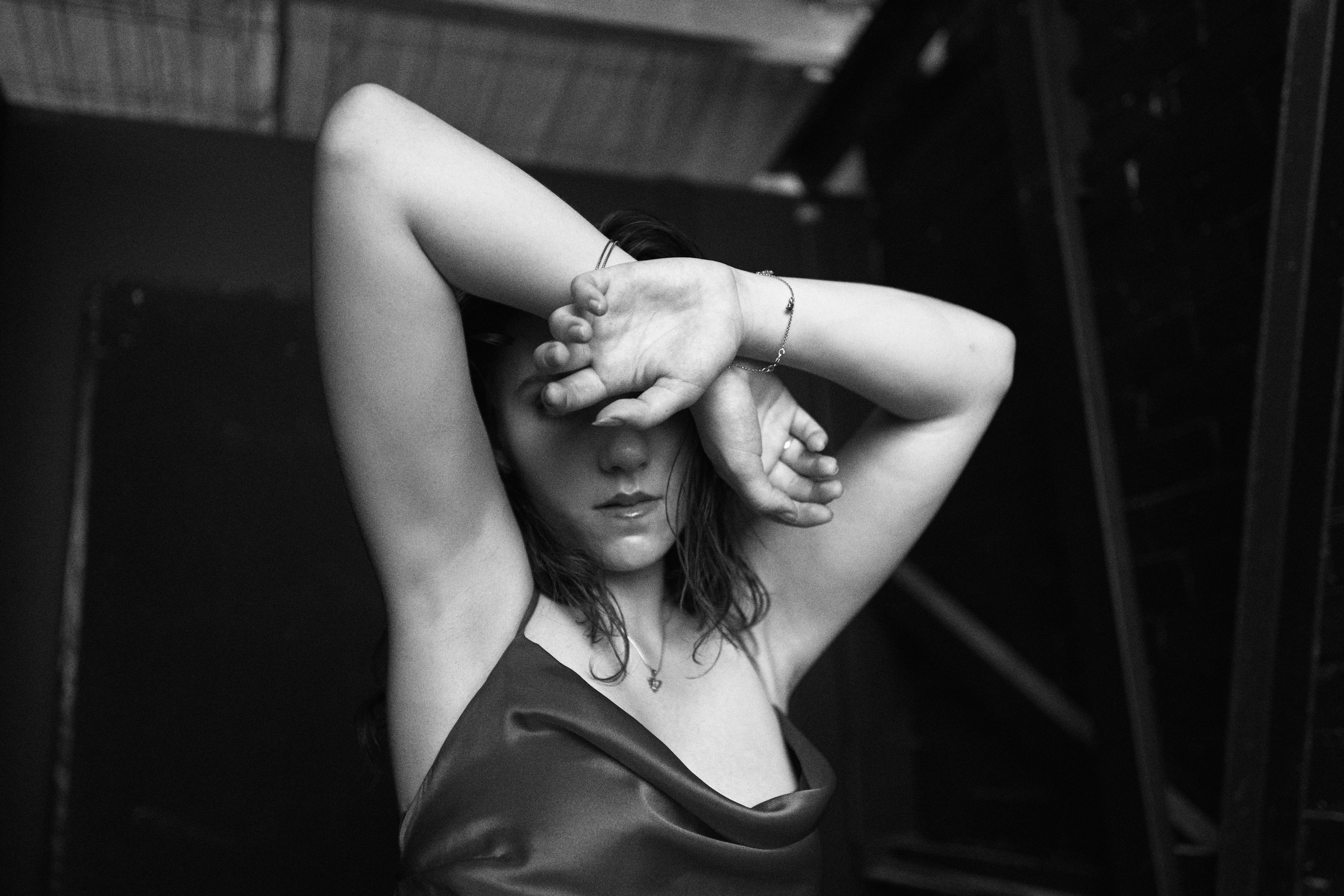 Monochrome portrait of a woman with hands covering her face, wearing a red satin top against a dark background. Intimate and artistic.